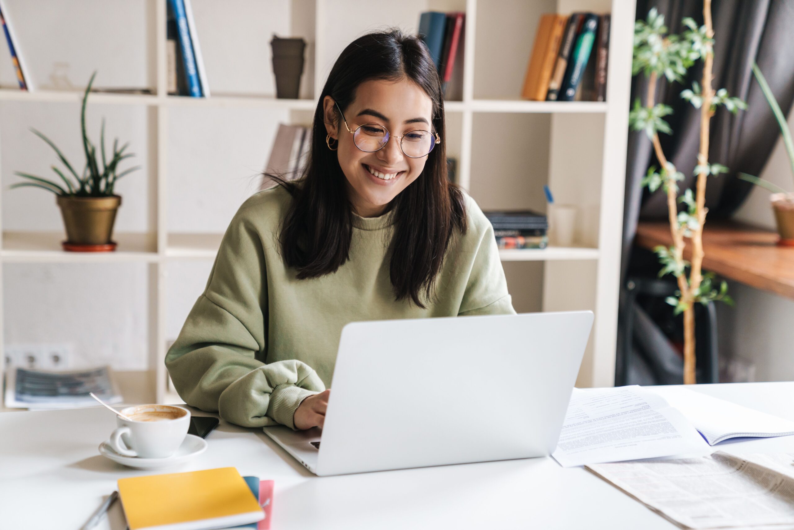 Young woman smiling while working on a laptop at a desk, with coffee and notebooks nearby, in a bright, organized study space.