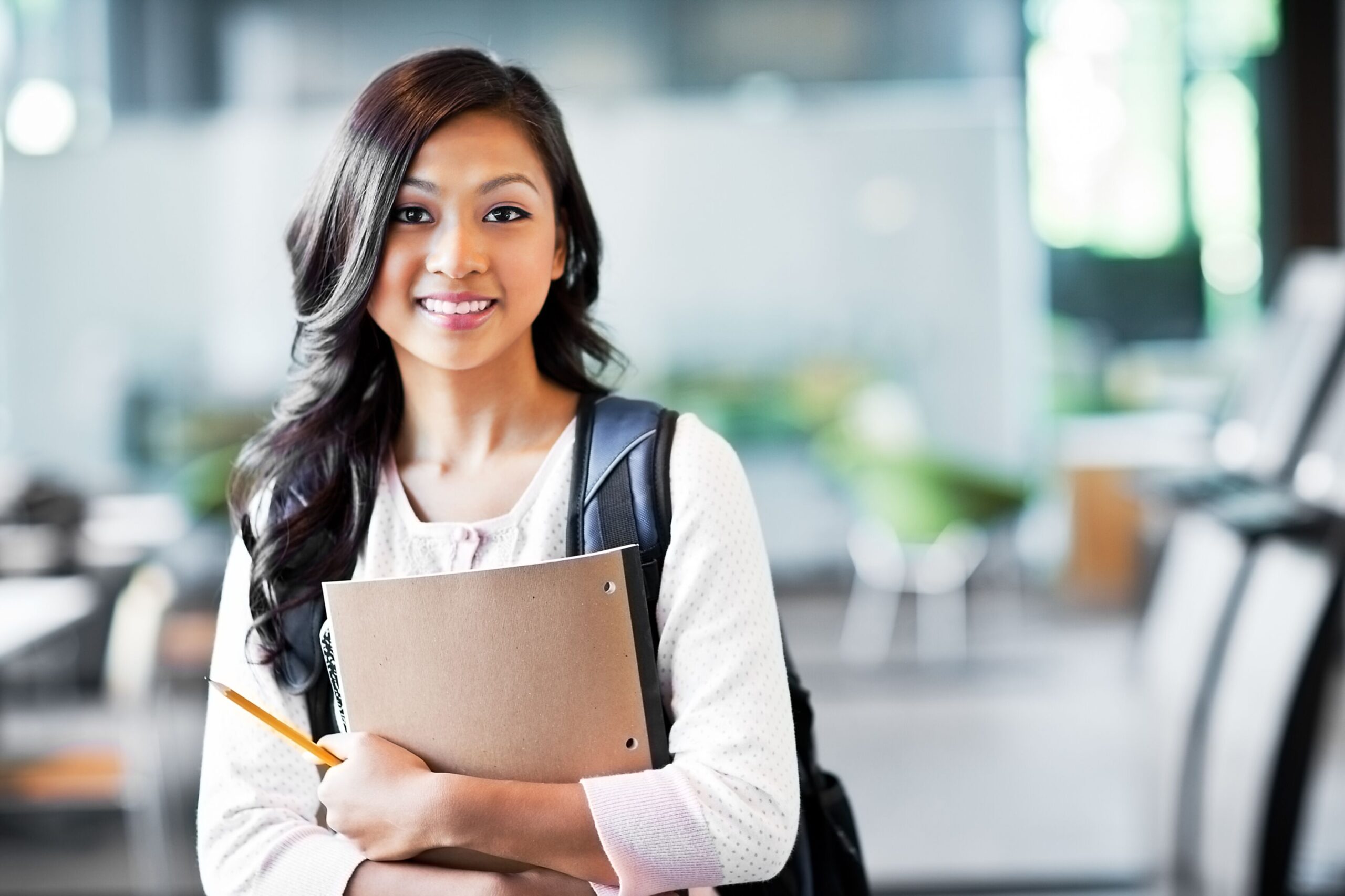 Confident student smiling while holding notebooks and a pencil in a modern educational environment, showcasing engagement and readiness to learn.